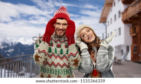 christmas, winter clothes and holidays concept - happy couple in ugly sweaters, knitted hats and mittens over ski resort in austrian alps mountains background