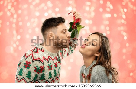 christmas, people and holiday traditions concept - portrait of happy couple in ugly sweaters kissing under mistletoe over festive lights on pink coral background