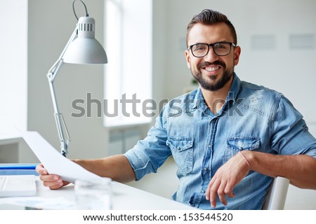 Smiling businessman in denim shirt and eyeglasses looking at camera in office Royalty-Free Stock Photo #153544520