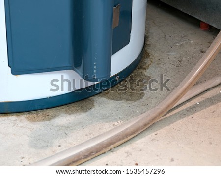 Water underneath a water heater shows the beginning of a leak, also shown by the signs of rust around the bottom rim, with selective focus on the water, and dirty hoses blurred in the foreground Royalty-Free Stock Photo #1535457296