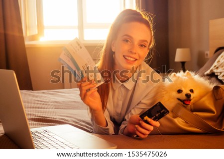 happy redhaired ginger woman lying down in bed with her cute white fluffy dog spitz in travel bag carrying and showing flyght tickets and credit card sundown light evening background