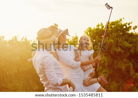 family, leisure and technology concept - happy mother, father and daughters having picnic and taking picture by smartphone on selfie stick at summer park. Sunset background