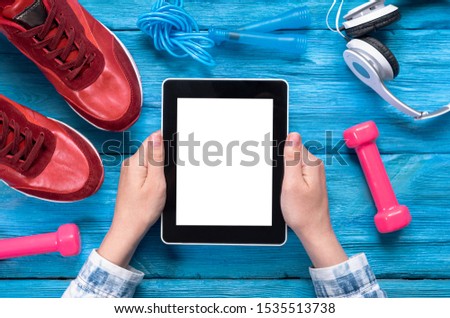 Tablet computer with black screen in female hand, red sneakers, jumping rope, dumbbells and headphones on blue wooden background.
