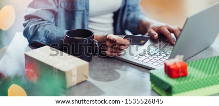 Woman wearing blue denim shirt sitting at the table by the Christmas tree with a credit card in her hand and shopping online on laptop
