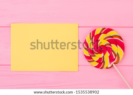 Spiral lollipop and blank paper sheet. Large multicolored lollipop and yellow paper card on pink wooden background.