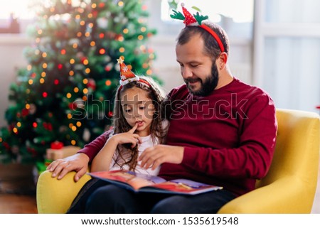 Man with beard wearing red sweater and his little daughter with dark hair reading Christmas fairy tales