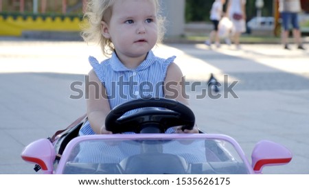 little girl in a blue dress rides a pink baby car in a park. Happy childhood concept. Caricature of an adult woman driving a car