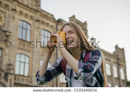 Young woman tourist taking photo on retro camera. Portrait of professional photographer holding yellow camera. Happy hipster traveler with backpack walking on street, laughing