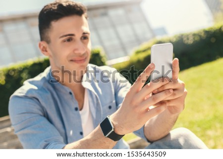 Stylish young man wearing digital watch sitting on concrete stairs outdoors in the urban park using application on smartphone close-up smiling happy blurred background
