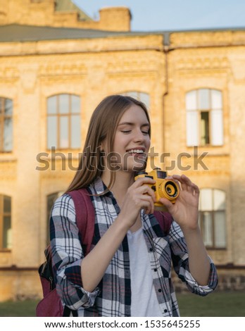 Happy hipster traveler with backpack walking on street, laughing. Young woman tourist taking photo on retro camera. Portrait of professional photographer holding yellow camera  