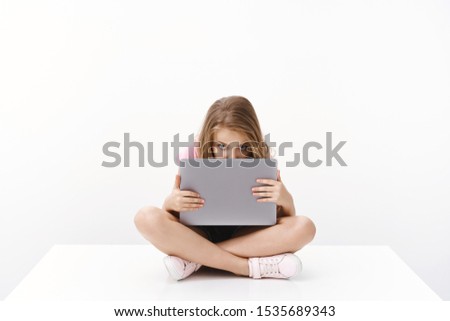 Cute cheerful charismatic blond young kid little girl student, sit on floor, hiding face behind laptop screen, stare camera bored, peeking, unwilling do homework wanna watch cartoons, play computer