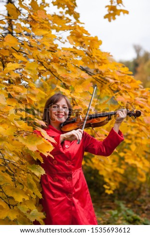 attractive young violinist in red wear plays violin among yellow autumn leaves of maple tree and smiles