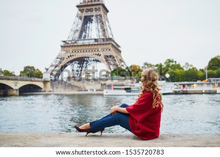 Young woman with long blond curly hair in Paris, France. Beautiful tourist in red coat near the Eiffel tower, on the Seine embankment