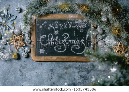 Christmas hand lettering Merry Christmas on vintage chalkboard with fir tree, golden stars, toys and lighted garland over blue texture background. Flat lay, space