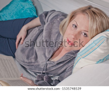 Beautiful blonde pregnant woman lying on the sofa and reading book, looking for information about pregnancy