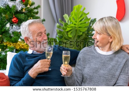 Senior couple man and woman with champagne and chatting, sitting on sofa with Christmas tree and decoration in background