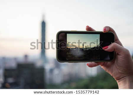 Asian woman hand with red nails holding a smartphone while taking a photo of Taipei cityscape during sunset time