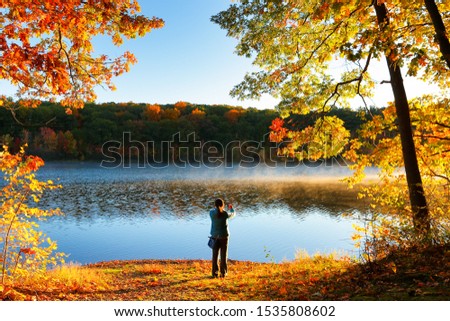 Beautiful Fall Foliage of New England at Sunrise, Boston Massachusetts. Photo shows a tourist using smart phone taking picture in front of lake.