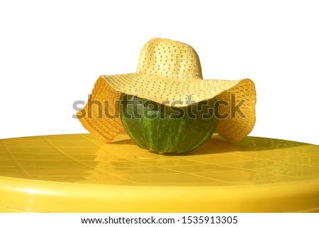   A green pumpkin in a bright yellow straw hat lies on a plastic yellow table outdoors, the main attribute of the fall Halloween holiday.                             