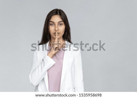 Young adult indian businesswoman in white formal wear standing isolated on grey background with copy space. Woman looking at camera and covering lips with finger, making silent sign, keep secret