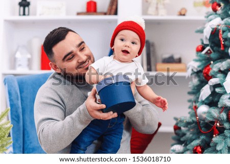 Young father sits in comfortable chair holding amusing toddler and gives gift box to him in the wonderfully decorated room to celebrate christmas.