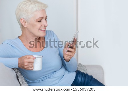 Attractive old business woman drinking coffee and using smartphone, sitting at sofa at home, copy space