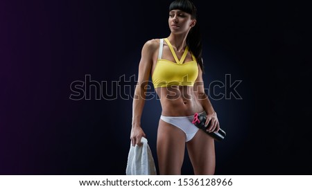 Female with perfect abdomen muscles on dark background with copyspace. Protein shake bottle and white towel in her hands.