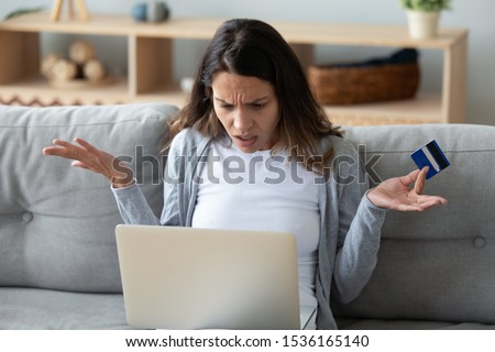 Irritated young mixed race girl sitting with computer and credit card on couch in living room, can not make purchase, not enough money. Unhappy client dissatisfied with online payment banking service. Royalty-Free Stock Photo #1536165140