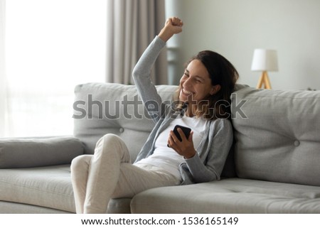 Hilarious euphoric millennial girl getting good news notification via mobile phone, making yes gesture, celebrating success, online lottery win, cheap flight tickets purchase, sitting on couch. Royalty-Free Stock Photo #1536165149