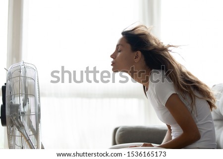 Side view young mixed race woman sitting on couch in front of ventilator indoors. Millennial girl suffering from hot summer weather or high temperature at home, cooling herself with air conditioner. Royalty-Free Stock Photo #1536165173