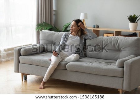 Full length positive dreamy young biracial girl leaning on comfortable couch, holding mobile phone in hands, looking at window, thinking of future date, meeting with friends or enjoying calm moment. Royalty-Free Stock Photo #1536165188