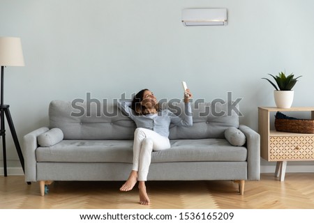 Front view full length joyful young mixed race woman relaxing on cozy couch in living room, holding remote controller, turning on cooler system air conditioner, setting comfortable temperature. Royalty-Free Stock Photo #1536165209