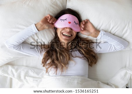 Above top view head shot happy millennial mixed race healthy woman lying in bed with sleeping mask, waking up after sweet dreams. Smiling young relaxed lady feeling energetic in morning at home. Royalty-Free Stock Photo #1536165251