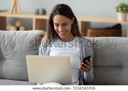 Head shot smiling young mixed race lady sitting on sofa at home with computer, shopping in internet, making purchases in online store, entering payment confirmation code from mobile phone message. Royalty-Free Stock Photo #1536165926