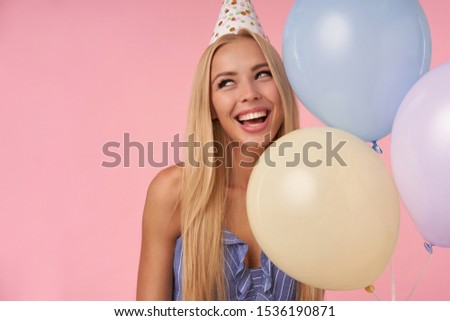 Close-up of charming young blonde female looking aside and smiling widely, posing in multicolored air balloons in blue summer dress and birthday cap, isolated over pink background