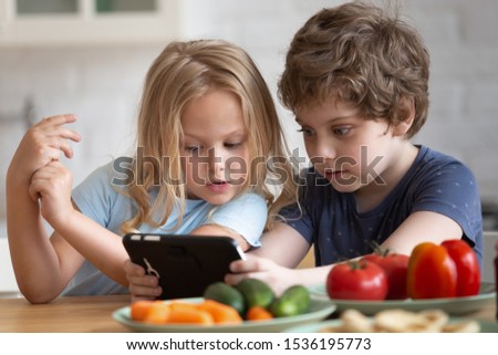 Curious little girl and boy using phone, sitting at table in kitchen, waiting for breakfast, funny sister and brother or friends looking at smartphone screen, watching video, playing game at home