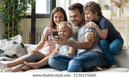 Happy parents with little children using phone together, sitting on couch at home, smiling father holding smartphone, taking selfie with kids, family watching video, having fun with cellphone