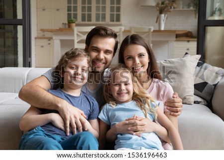 Head shot portrait happy family with two kids at home, smiling mother and father hugging with children, posing for picture in living room, young parents with little daughter and son looking at camera
