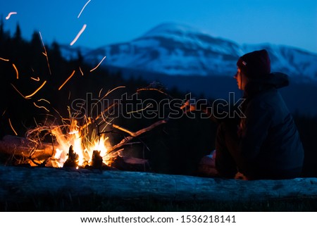 Girl relaxes near a bright campfire with sparks. Forest and snow-capped mountain in the background. Evening in the Carpathians. Royalty-Free Stock Photo #1536218141