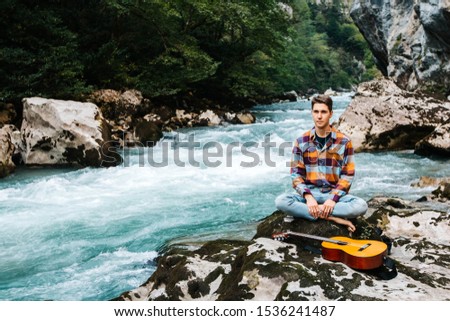 Man in a meditative position with guitar sitting on the bank of a mountain river on a background of rocks and forest. Concept of freedom relaxation. Place for text or advertising