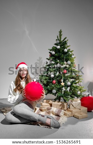 A young girl in a Christmas hat and a white knitted sweater with a large red ball in her hands sits on a plaid against the background of a Christmas tree. Christmas and new year concept.
