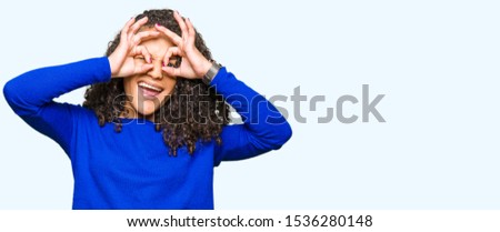 Young beautiful woman with curly hair wearing winter sweater doing ok gesture like binoculars sticking tongue out, eyes looking through fingers. Crazy expression.