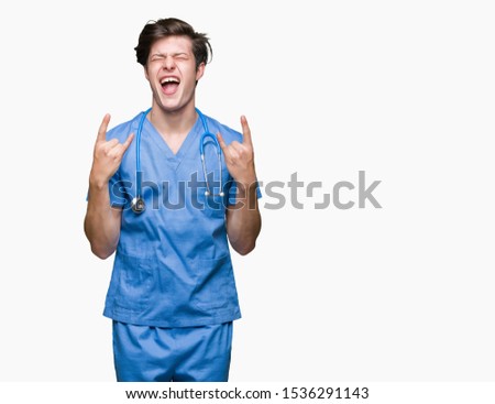 Young doctor wearing medical uniform over isolated background shouting with crazy expression doing rock symbol with hands up. Music star. Heavy concept.