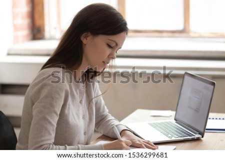 Concentrated caucasian girl sit at desk studying preparing for university session examination holding pen writing down in textbook, focused smart female student make notes do task using laptop indoors Royalty-Free Stock Photo #1536299681