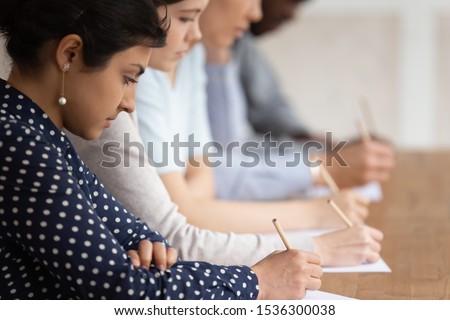 Multinational students seated at desk in row holding pencils writing on papers, take part in university exams, learning process or test of scholars knowledge skill in subject, higher education concept