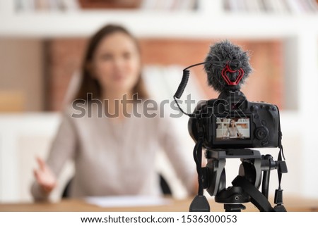 Focus on foreground camera screen show live recording video blog, confident millennial woman seated in front of cam filming educational video for online university course, share professional knowledge