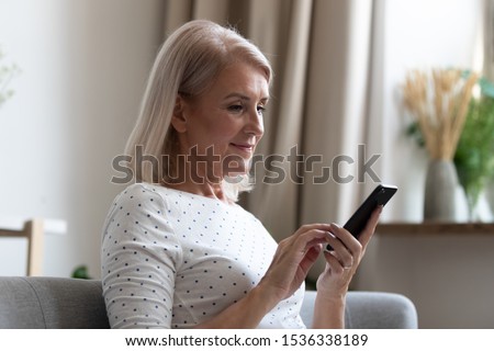 Happy middle aged mature woman enjoying using mobile apps texting typing messages sit on sofa, smiling old adult lady holding smartphone looking at cellphone screen browsing social media at home Royalty-Free Stock Photo #1536338189