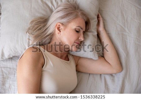 Peaceful healthy beautiful 50s mature woman lying asleep on comfortable pillow orthopedic mattress sleeping well in cozy bed alone, calm serene old female resting in bedroom, close up top view