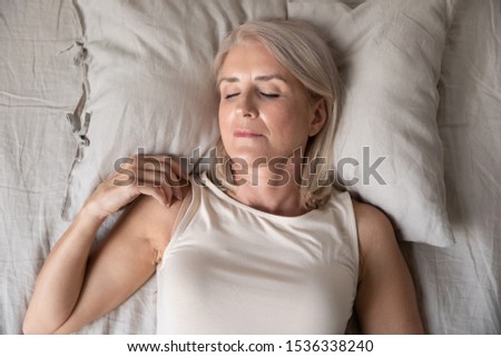 Healthy serene mature middle aged woman sleeping alone in comfortable cozy bed on soft pillow linen orthopedic mattress, calm old lady enjoy peaceful sleep concept relax at home, close up top view