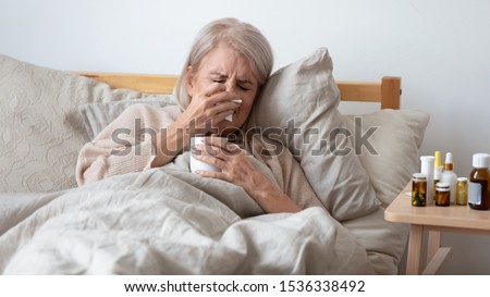 Older mature upset ill woman holding handkerchief blowing running nose in bed at home, sick old senior lady got infectious influenza virus disease flu grippe symptoms sneezing caught cold concept Royalty-Free Stock Photo #1536338492